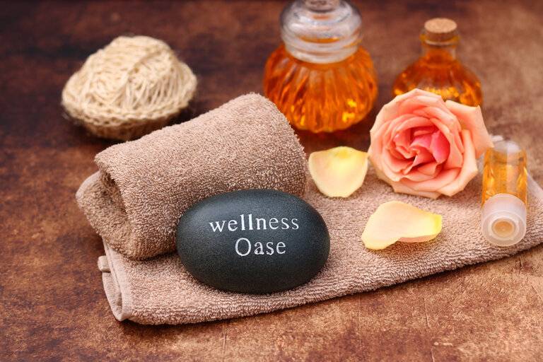 Spa-Komposition: Stein mit dem Text Wellness-Oase auf einem Handtuch mit Aromaöl und Rosen.