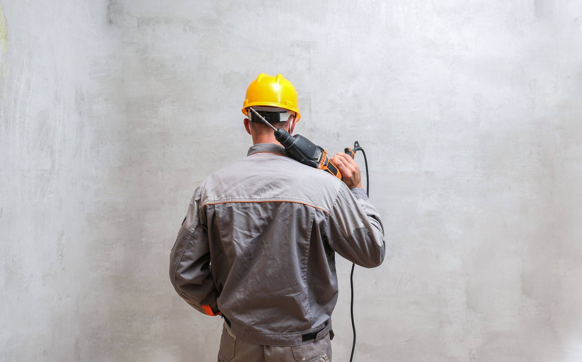 Handwerker mit Schutzhelm und Bohrmaschine steht vor Betonwand – symbolisiert Vorbereitung für Befestigung mit Betonverankerung